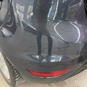 Kratzer und Dellen an der hinteren Stoßstange eines dunklen Autos in einer Werkstatt.