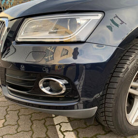 Frontstoßstange eines dunklen Autos mit Lackschäden und Scheinwerfer auf Parkplatz.